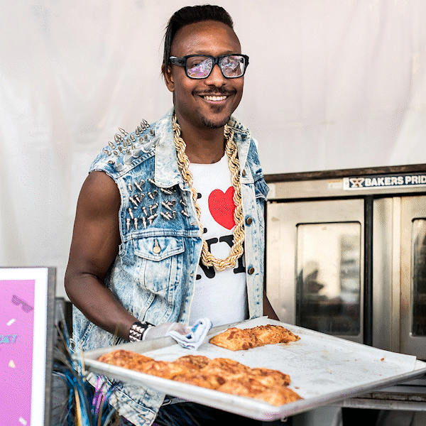 Gregory Gourdet in a denim vest, gold chains and I heart NY teeshirt pulling pastries out of an oven at the East Coast vs West Coast Feast event. A cartoon heart pulses from his heart on his t-shirt