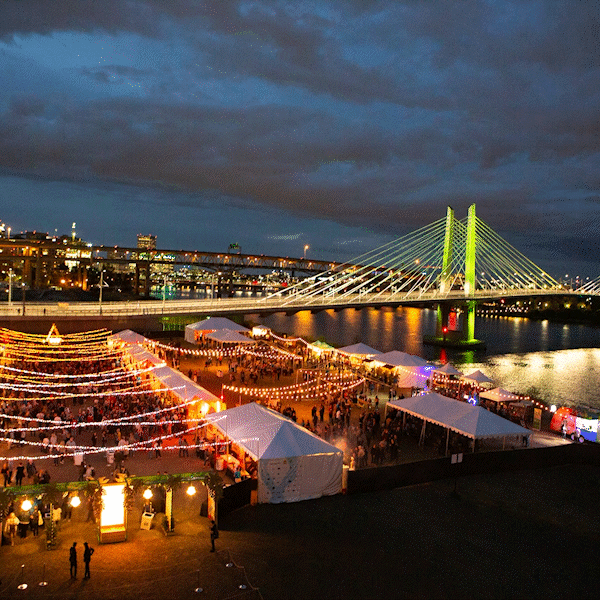 Rooftop photo of the the Feast Night Market event at night with tents lit up and a view of the Tillicum bridge and the Willamette River. A cartoon image of a flying shrimp rolls through the sky