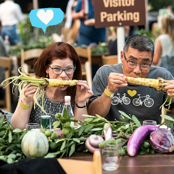 A man and woman sitting at a table biting into corn on the cob with a gif of a blue heart pulsing over the woman's head