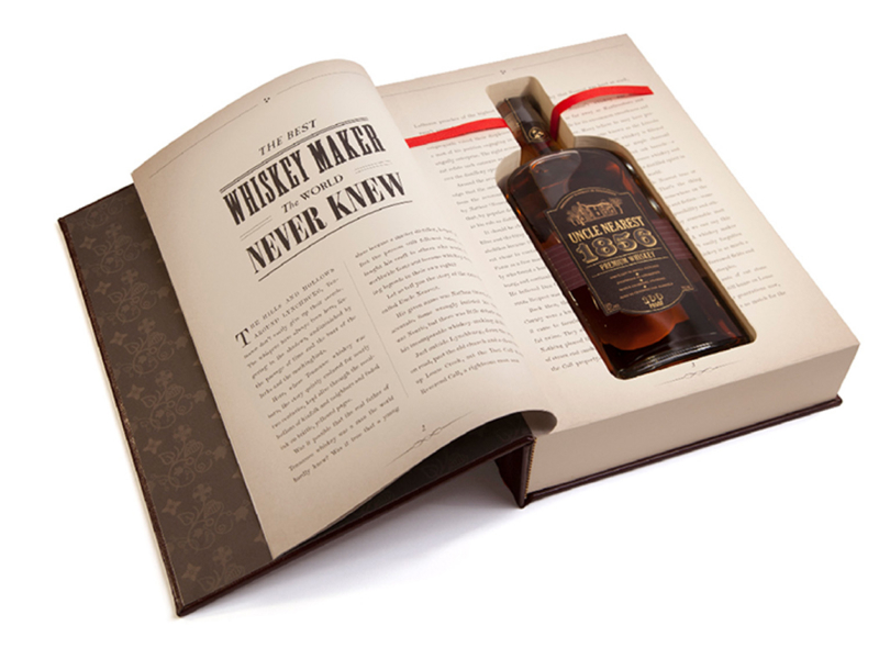 Uncle Nearest Launch Book – Open with Bottle showing, - the seonc page of the book is open and shows that is a full sized whiskey bottle cut out and fitted inside the book. There is a red ribbon tucked behind the whiksey bottle neck to pull the bottle out of the book. On the opposite page is reads 